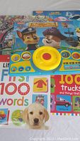 Five kids books arranged closely, showing colorful covers including Paw Patrol 'Pups and the Pirate Treasure' with attached blue and yellow sound panel, and other books featuring first 100 words and trucks.