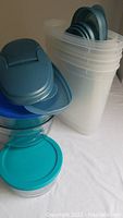 Stack of various white plastic containers with blue lids and clear glass containers with turquoise lids on a white surface, needing cleaning.