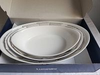 Three white oval glass baking dishes nested inside each other in the box. New condition, no visible marks or defects.