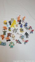 Overview photo of assortment of about 30 miniature toy figures on white background, showing variety of colors and shapes including animals, robots, and humanoid forms.