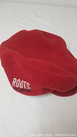 Close-up of red fleece hat showing embroidered Roots Athletics logo on side.