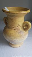 Yellow clay vase with decorative tarnish, scroll handles on the sides, and a zigzag pattern near the neck.