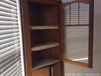 Interior view of corner hutch cabinet with glass door open, showing two triangular shelves with fabric liners and lower enclosed storage.