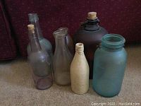 Group of seven assorted glass bottles of various colors and sizes, some with cork stoppers, placed on floor in front of couch.