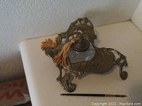 Brass ornate inkwell holder with detailed scrollwork shown on a white surface. A clear glass inkwell fits inside the holder. A gold-colored pen with a tassel lies adjacent to the holder.