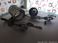 Photo of two handheld egg beaters with crank handles and large vintage flour sifter with crank handle showing some rust and wear, in front of wallpaper with kitchen utensil design.