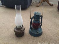 Photo shows two kerosene lanterns. One is a blue metal framed lantern with red glass inside and a carrying handle. The other is a bronze metal base lantern with a tall white frosted glass chimney. Both are placed on a beige carpeted floor.