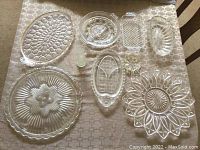 Nine clear glass serving plates and dishes laid on a tablecloth showing different shapes, styles, and designs.