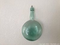 Front view of old green hanging bottle showing round body, long neck, and hook on top against a white textured wall.
