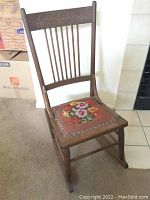 Full view of the wooden rocking chair showing the slatted back and carved top rail with embroidered seat.