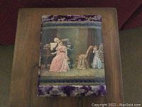 Front cover of the photo album showing a soft purple textured fabric with a vintage Victorian scene in the center, trimmed with lace and a metal clasp on the right side.