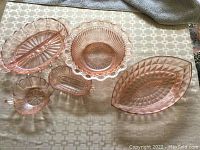 Six pink glass dishes arranged to display various shapes and patterns including oval, round, flower-shaped, and leaf-shaped forms on a patterned tablecloth.