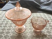 Pink depression glass covered candy dish and creamer featuring embossed rose patterns on a fabric surface.