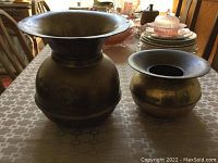 Two metal spittoons side by side on table. Larger spittoon has a wide flared rim with bulbous body; smaller spittoon has similar shape but smaller size.