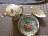 Top view showing the tea pot with floral design on lid and body, white gravy boat, and decorative plate with classical romantic scene and pierced gold decorations.