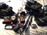 Displays numerous vintage box cameras with leather cases, a silver compact camera, Canon charger, tripod and binoculars on a bed surface.