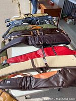 Photo of multiple soft rifle and hunting bow cases and carriers on tables outdoors, showing various colors and materials, pre-owned with signs of wear.