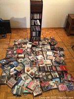 Wide view of tall wooden CD rack with multiple shelves filled with CDs, surrounded by a large spread of loose CDs on the floor.