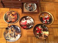 Six collector plates arranged on the floor showing various Christmas and winter scenes including Santa Claus and snowy streetscape.