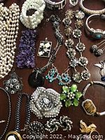 Photo displaying a mix of costume jewelry on a brown surface, including necklaces with pearls and chains, amethyst stone bracelets, turquoise earrings, and rhinestone brooches.