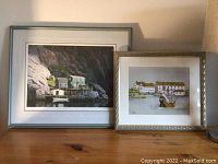 Two framed art prints set on a table against a wall, the larger showing a rocky coastal building scene and the smaller a harbor with white buildings and ship.