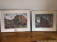 Two framed art prints side by side on floor; left print 'Dunlan's Open Kitchen' featuring streetcar, right print 'The Gooderham Building' cityscape