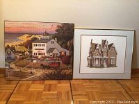 Photo showing two framed art pieces on the floor: a large puzzle art of coastal scenery and a framed architectural house illustration.