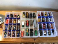 Photo showing multiple Hot Wheels cars organized in a plastic storage case with clear compartments and blue dividers on a wooden surface.