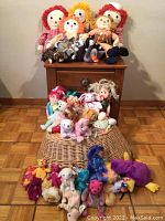 Raggedy Ann and Andy dolls sitting on wooden furniture, surrounded by various Ty Beanie Babies and plush toys on and around a wicker basket on the floor.