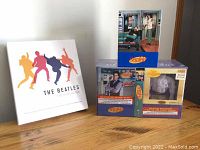 Photo showing Beatles box set with colorful silhouettes and the largest Seinfeld box set with seasons 5 & 6 figurine on a wood surface.