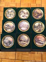 Nine Thomas Kinkade collector plates arranged on a floor, each with winter scenic artwork and gold decorative rim.
