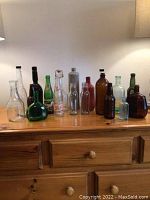 Full lineup of 13 vintage glass bottles of assorted styles and colors displayed on wooden surface under lamp lighting.