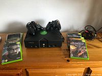 Front view of Xbox console with two controllers on top and 6 game cases around it on a wooden surface.