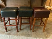 Three square top bar stools, two black and one brown, wood legs, standing side-by-side showing full height and seat dimensions.
