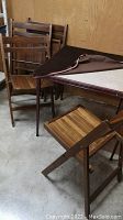 Vintage folding card table with dark brown cover partially lifted to show beige playing surface, with 4 matching wooden folding chairs behind and next to it.