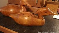 Two wood carved duck figurines displayed on a black surface in front of a wooden cabinet. The larger duck features carved eyes, and the smaller duck has inset eyes.