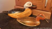 Two carved wooden duck decoys shown side by side on a dark countertop with a kitchen cabinet background.