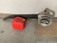 Full view of the leaf blower with black nozzle and gray motor casing with red trigger, alongside red fuel can labeled BLOWER.