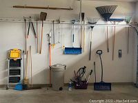 Wide view of garden and automotive tools hanging and leaning against a wall including rakes, shovels, pruners, brooms, folding ladder, and garbage can.