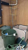 Photo showing one large green outdoor waste can with lid, two collapsible beige and green garden bins, and green gardening hand tools including a hand rake and cultivator.