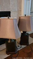 Pair of table lamps with beige fabric shades and metal bases with wooden bottoms seen on a floor in front of tile wall