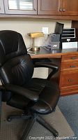 Black executive style office chair on five caster wheels with clearly visible padded armrests and cushioned seat and backrest. Chair rests on a clear textured floor mat. Behind the chair, a brass-colored desk lamp and transparent plastic paper sorter with multiple compartments are visible on a wooden desk.