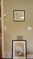 Full view of two framed paintings hung on a wall, one small above and one larger below the air vent