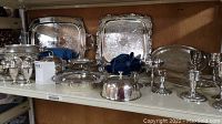 Wide view of multiple silverplate trays, servers, covered butter dish, salt and pepper shakers, cream and sugar cups, and candle holders on shelf.