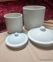 Two white ceramic canisters with matching lids displayed on beige fabric with a red patterned background. Both canisters feature glossy finish and rounded lids with knob handles evident.