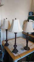 Photo showing all three lamps on a table with white tile surface, two taller lamps measuring 30 inches and one shorter at 28.5 inches, all with white shades and bronze-like ornate bases.