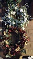 Photo of two wreaths stacked vertically: top wreath white and green floral with soft white flowers, bottom wreath with deep red and cream flowers and green leaves.