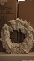 Full view of a circular white ceramic wreath decorated with carved angels and flowers, placed against cardboard boxes.