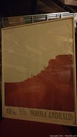 Framed signed print titled "Pueblos" by Norma Andraud visible through glass with red and cream stylized pueblo design.