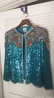 Front view of turquoise sequin blazer with colorful beaded paisley pattern on shoulders and chest area hanging on wooden hanger.